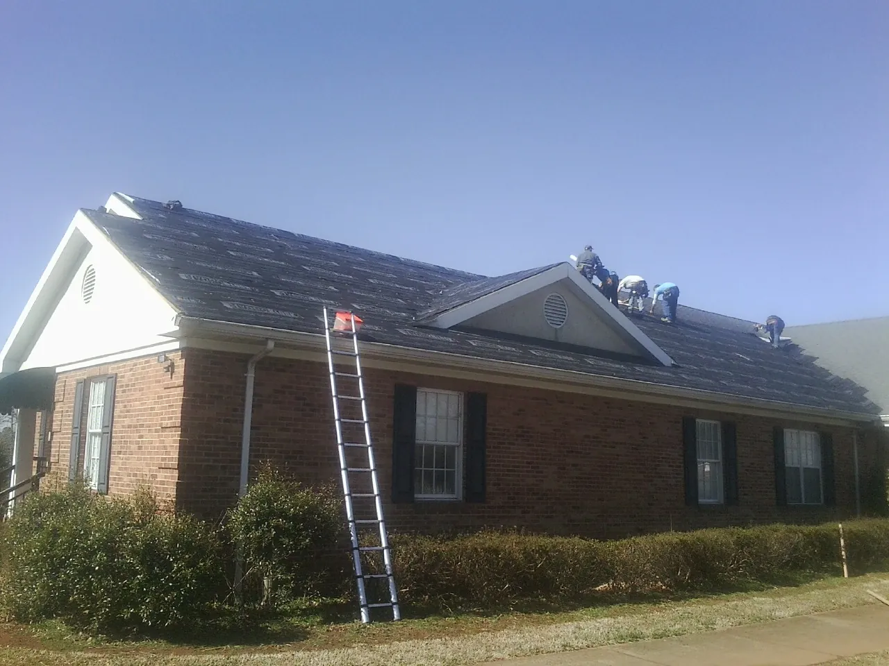 Crew tearing shingles off a brick ranch roof with a ladder set — Metal Roof Repair work by our crew in Mill Neck