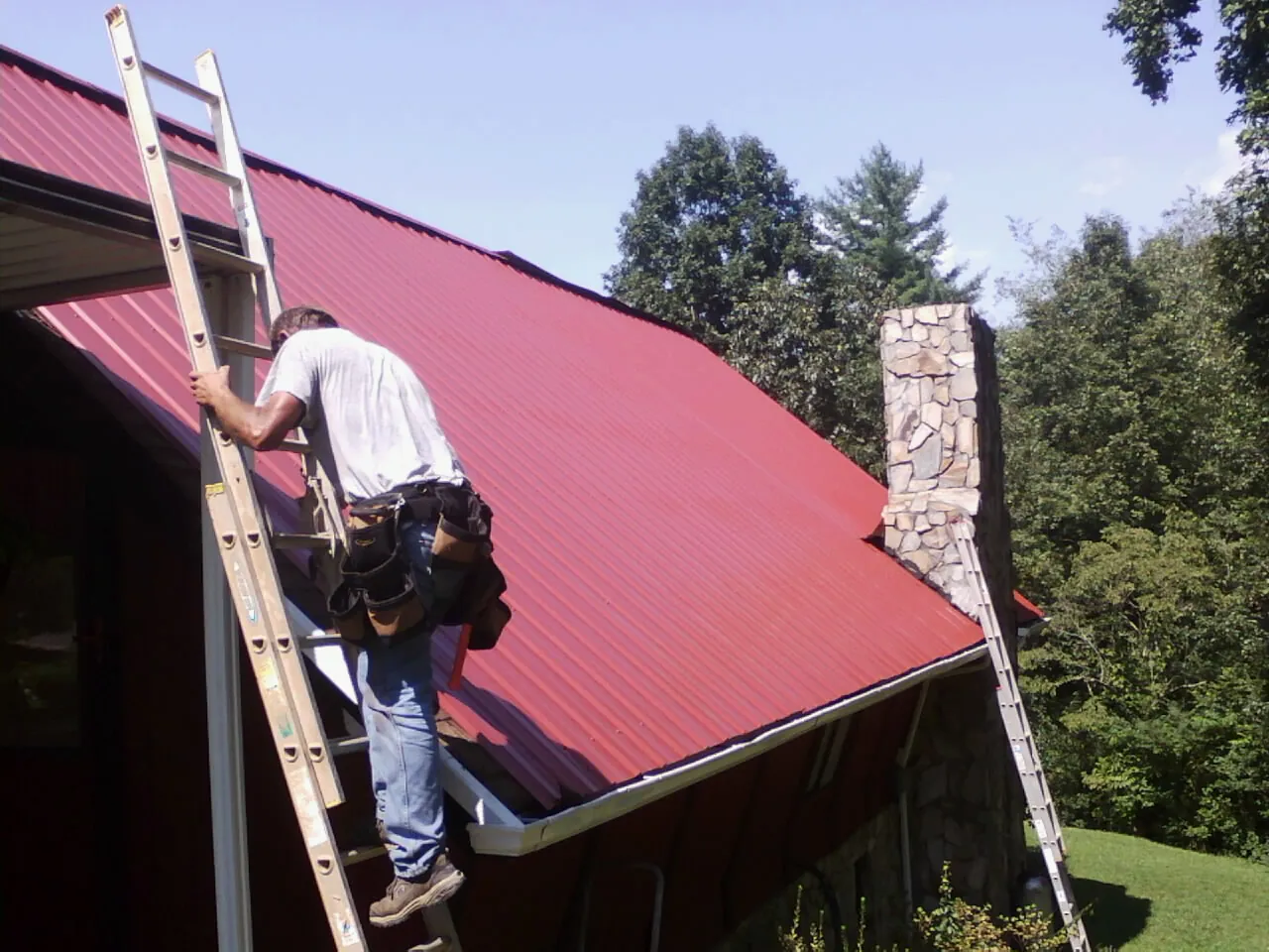 Roofer on a ladder beside a red metal roof and stone chimney — Soffit & Fascia Repair by Roofing Brothers in Mill Neck