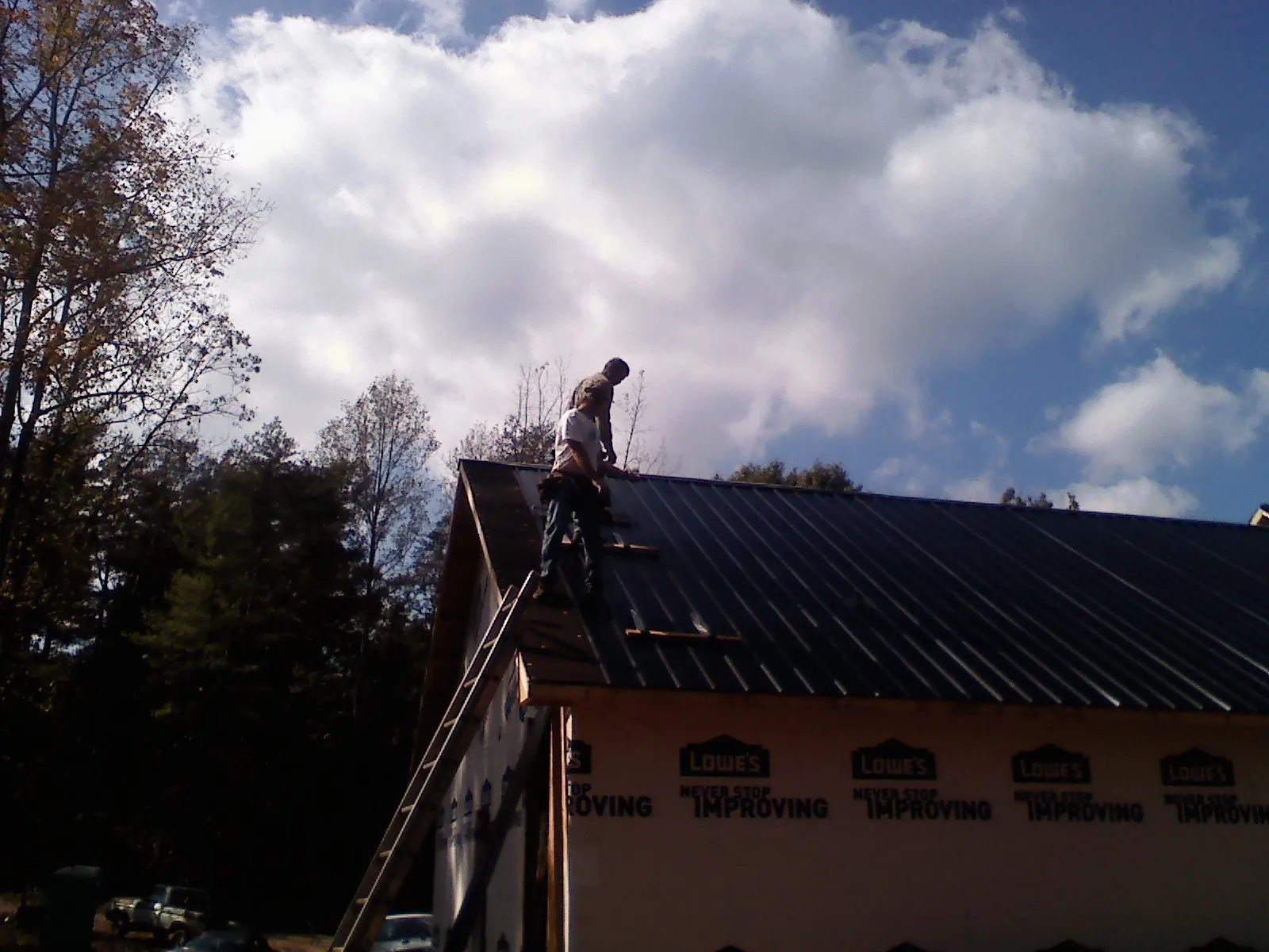 Roofer fastening dark metal panels high against a cloudy sky, from a Roof Inspection job in Mill Neck